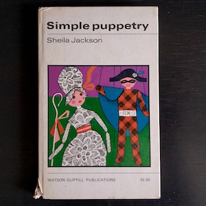 May include: A book titled "Simple Puppetry" by Sheila Jackson. The cover features a colorful illustration of two puppets, a female puppet wearing a white lace dress and a male puppet wearing a red and black checkered costume.