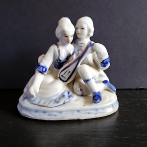May include: A porcelain figurine depicting a seated couple in period clothing. The figures are primarily white with blue and gold accents. The man is holding a stringed instrument. The base is oval-shaped.