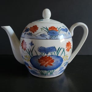 May include: A white ceramic teapot with a lid and handle, decorated with a floral design. The design features orange and red flowers, blue lily pads, and green stems. The teapot has a curved spout and a rounded body.