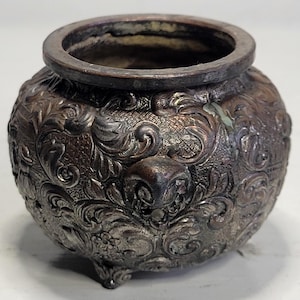 May include: A small, antique silver-colored pot with ornate floral and scrollwork detailing. The pot has a rounded body, a wide opening, and three small feet. The surface shows signs of age and wear, adding to its vintage charm. A decorative piece.