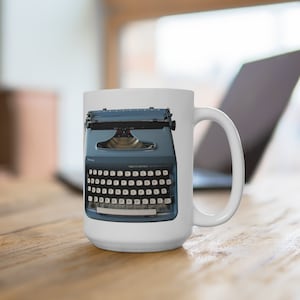 May include: A white ceramic mug with a blue typewriter printed on it. The typewriter has a white keyboard with black lettering and the words "Remington" and "Model 5" printed on it.