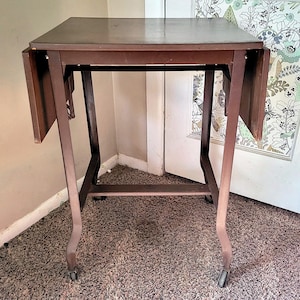 May include: A dark brown, antique-style side table with drop leaves. The table has a rectangular top, curved legs, and small wheels. The table is made of wood and has a simple, elegant design. The table is approximately 28 inches tall.