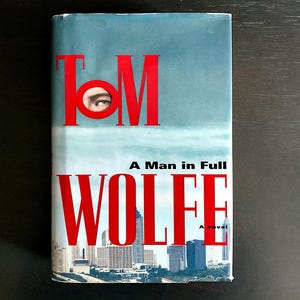 May include: A book cover with the title "TOM WOLFE" in red letters. The title "A Man in Full" is written in black letters below the author's name. The cover features a cityscape with a close-up of an eye peeking through a hole in the letter "O" in the author's name.