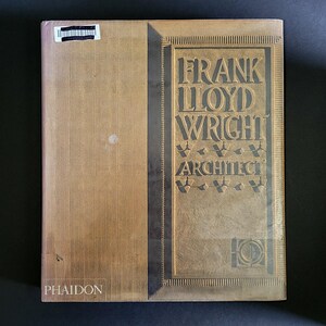 May include: A hardcover book titled Frank Lloyd Wright Architect published by Phaidon. The cover features a textured tan and gold surface with vertical lines on the left and embossed lettering on the right. A small black barcode sticker is visible.