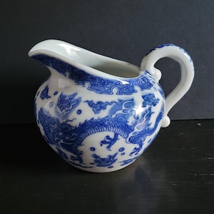 May include: A small, white ceramic creamer with a blue dragon and cloud pattern. The creamer has a curved handle and a spout for easy pouring. The design is a classic Asian-inspired motif.