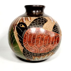 May include: A round, dark-necked ceramic vase featuring a detailed, hand-carved relief of a sea turtle with a textured, reddish-brown shell. The design includes black, green, and tan accents, with carved leaf patterns surrounding the turtle.