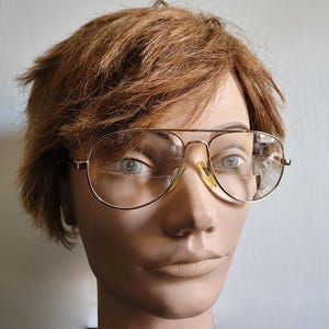 May include: Gold-toned metal aviator-style eyeglasses with a double bridge design and clear lenses. The frames feature adjustable nose pads and thin temples, worn on a mannequin head with short, textured, light brown hair.