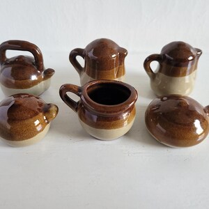 May include: A collection of six small, brown and beige ceramic pots and pitchers. The pieces vary in shape, including a kettle with a handle, small pitchers with handles, and a round pot with a small handle.