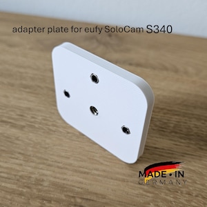 May include: A white adapter plate for the eufy SoloCam S340, with four circular mounting holes. The plate is made in Germany and rests on a wooden surface. The text "adapter plate for eufy SoloCam S340" is visible.