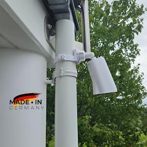 Könnte beinhalten: Eine weiße Überwachungskamera, die mit einer Halterung an einem weißen Rohr befestigt ist. Die Kamera ist nach unten geneigt. Das Bild enthält den Text "MADE IN GERMANY" mit den Farben der deutschen Flagge.