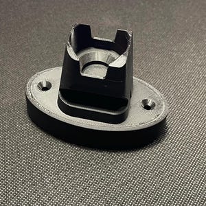 May include: A black 3D printed wall mount with a rectangular opening for a device. The mount has two small holes on the side for mounting.