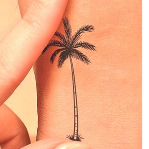May include: A black palm tree tattoo on a person's arm. The tattoo is simple and minimalist, with a single palm tree trunk and leaves.