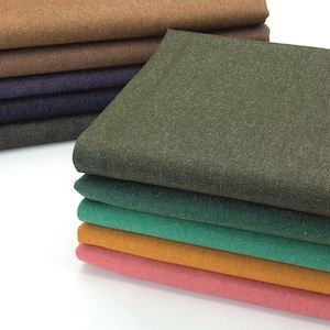 May include: A stack of folded fabric pieces in various solid colors including olive green, teal, mustard yellow, pink, dark gray, navy blue, purple, and shades of brown. Each piece features a visible woven texture and a consistent rectangular shape.