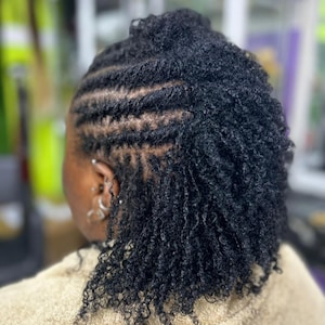 Op de afbeelding: Een persoon met zwarte dreadlocks die zijn haar heeft gestyled met een scheiding aan de zijkant van zijn hoofd. De dreadlocks zijn netjes gestyled en lijken vers te zijn.
