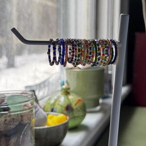 May include: A white metal ring holder with a variety of colourful beaded rings. The rings are made of small, colourful beads and are arranged in a row on the holder.