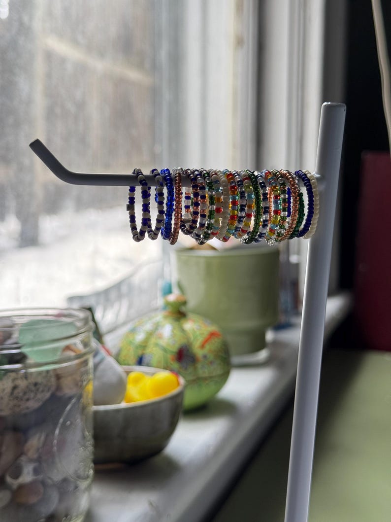 May include: A white metal ring holder with a variety of colourful beaded rings. The rings are made of small, colourful beads and are arranged in a row on the holder.
