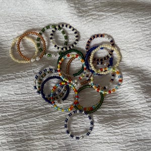 May include: A collection of colourful beaded bracelets in various shades of blue, green, white, and orange. The bracelets are made with small, round beads and are stacked on top of each other.