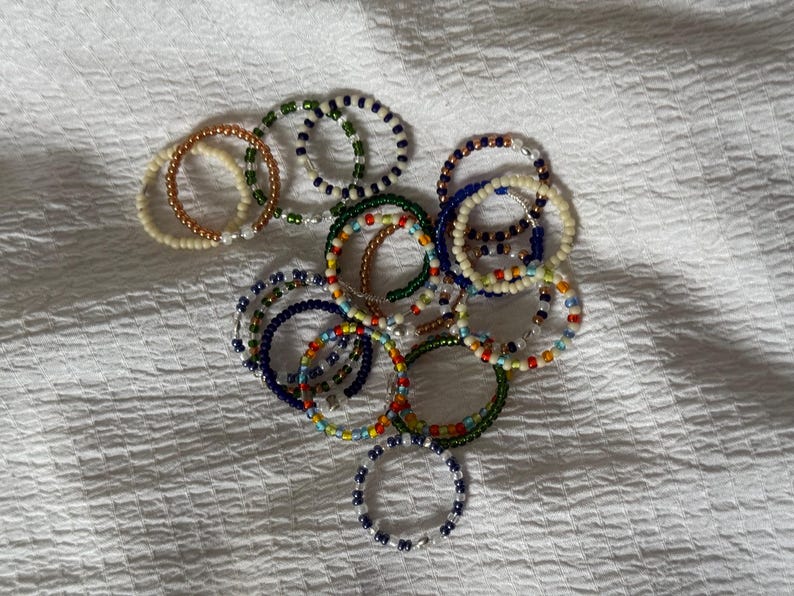 May include: A collection of colourful beaded bracelets in various shades of blue, green, white, and orange. The bracelets are made with small, round beads and are stacked on top of each other.