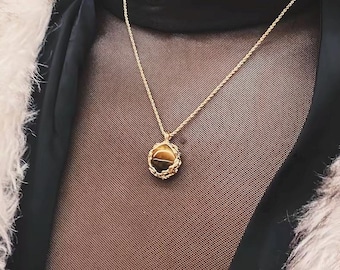 May include: A gold-colored necklace with a pendant featuring a brown and gold stone. The pendant is oval-shaped and surrounded by a decorative gold frame. The necklace is displayed against a dark background.
