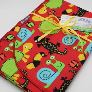 May include: Red fabric baby blanket with a colorful pattern of snails, turtles, and lizards. The blanket is tied with a yellow ribbon and has a small tag that reads "Kim Williamson Creations, Baby Blankets & Burp Cloths".