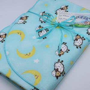 May include: A light blue baby blanket with a white sheep and crescent moon pattern. The blanket is tied with a blue ribbon and a tag that reads "Kim Williams Baby Wrap".