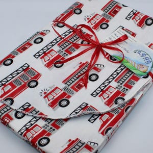 May include: A white baby blanket with a repeating pattern of red fire trucks. The blanket is tied with a red ribbon and has a tag attached. The fire trucks have black tires and ladders. The blanket is folded.