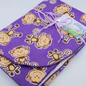 May include: Purple fleece baby blanket with a brown and pink monkey pattern. The blanket is folded and tied with a purple ribbon.