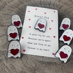 Puede incluir: Un conjunto de pines blancos con forma de fantasma y detalles de corazón rojo, dispuestos alrededor de una tarjeta. La tarjeta dice "Ghost Hug" e incluye un mensaje sobre el recuerdo. Los pines y la tarjeta están sobre una superficie de madera.