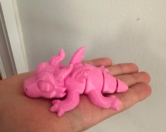 May include: A pink 3D printed dragon figurine with detailed scales and wings. The dragon is lying on its side with its head resting on its front paws.