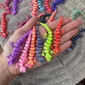 Könnte beinhalten: Eine Sammlung farbenfroher, beweglicher Eidechsenfiguren in verschiedenen Lila-, Rosa-, Orange-, Blau- und Grüntönen. Die Eidechsen sind auf einer Holzoberfläche angeordnet, einige werden in einer Hand gehalten. Die Figuren haben detaillierte, segmentierte Körper und kleine Beine.