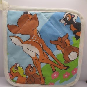 May include: A square quilted pot holder featuring a printed scene of a brown deer, a black and white skunk, and a brown rabbit on a light blue background with green grass and pink flowers. The edges are finished with a cream-colored fabric trim.