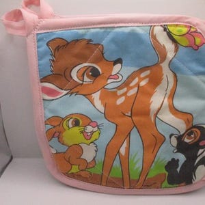May include: A square quilted pot holder with a light pink fabric border and a hanging loop. The center features a colorful illustration of a brown fawn, a small yellow rabbit, and a black and white skunk against a light blue sky background.
