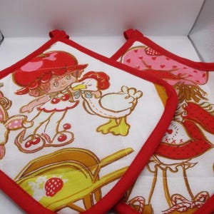 May include: Two square fabric pot holders with a white background and a red fabric border. The print features a cartoon character with red hair, a white dress with cherry accents, a white goose, a yellow wheelbarrow, and a rabbit.