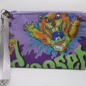 May include: A rectangular purple fabric zipper pouch featuring a graphic of an orange, snarling creature with green slime. The pouch includes a detachable gray and white patterned wrist strap attached to a silver metal clasp on the side.