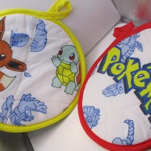 May include: Two round, white fabric pot holders with colorful Pokemon characters. One pot holder has a yellow border and features a Squirtle, a Bulbasaur, and a Pikachu. The other pot holder has a red border and features the word "Pokemon" in yellow letters.