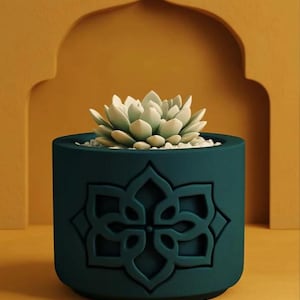 May include: A teal cylindrical plant pot with a raised floral pattern. It contains a light green succulent and small white pebbles. The background is mustard yellow, with an arch-shaped design. The pot is suitable for indoor plants.