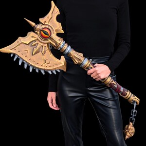 May include: A large, ornate battle axe with a gold and silver finish. The axe head features a stylized eye and jagged teeth along the blade. The handle is detailed with intricate designs and a chain with a skull pendant.