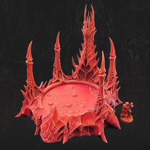 May include: A red miniature terrain piece featuring a circular platform surrounded by five tall, jagged, organic-looking spires. A small, hooded figure stands to the right of the structure, which has a textured, bubbling surface on the base.