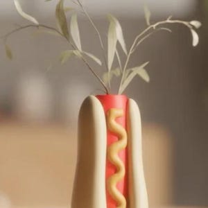 May include: A whimsical ceramic vase shaped like a hot dog, complete with a red interior, beige bun, and a yellow mustard design. The vase holds delicate green plant stems, adding a touch of nature to the playful design. A unique decorative piece.