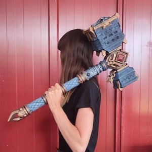 Puede incluir: Un gran martillo ornamentado con una combinación de colores azul y dorado. El martillo presenta detalles intrincados, incluyendo runas y patrones geométricos. El mango es azul claro con un diseño texturizado. El martillo está siendo sostenido por una persona.