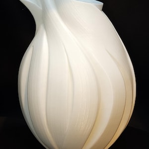 May include: A white ceramic vase with a unique, organic shape resembling a flower bud. The vase has a smooth, textured surface and a wide opening at the top.
