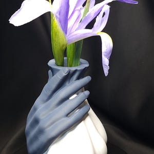 Könnte beinhalten: Eine einzigartige Vase mit einem weißen, bauchigen Sockel und einem grauen Handdesign, das die Oberseite umfasst. Sie enthält eine violett-weiße Irisblume mit grünen Stielen, vor dunklem Hintergrund.