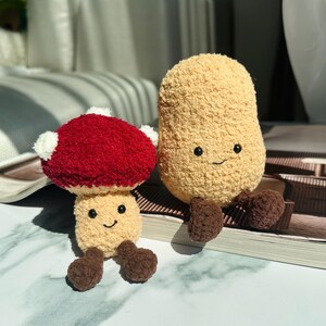Può includere: Due peluche: un fungo rosso e bianco e una figura beige a forma di fagiolo, entrambi con facce sorridenti e piedi marroni. Il fungo ha un cappello rosso e dettagli bianchi. Il giocattolo a forma di fagiolo ha una superficie strutturata.
