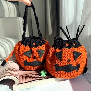 May include: Two orange crochet pumpkin bags with black accents and jack-o'-lantern faces. One bag has a black handle, and the other has black stems. The bags are sitting on a magazine.