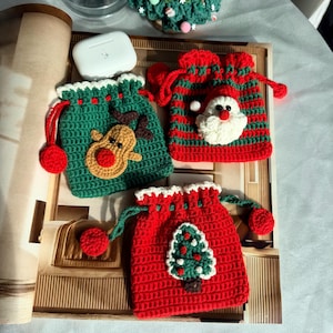 May include: Crocheted Christmas-themed pouches in green and red, featuring a reindeer, Santa Claus, and a Christmas tree design. Each pouch has a drawstring closure and a decorative pom-pom. The pouches are handmade.
