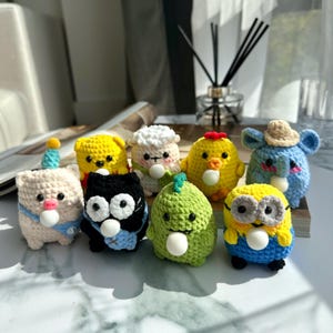 Könnte beinhalten: Acht gehäkelte Stofftiere in verschiedenen Farben und Designs, darunter ein Schwein, eine Katze, ein Dinosaurier, ein Küken, ein Schaf, ein Bär, ein Elefant und ein Minion. Jedes Tier hält einen weißen Ball im Maul.