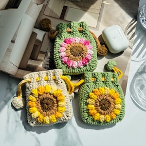 May include: Three crochet drawstring pouches with sunflower designs. The pouches are in shades of green, beige, and yellow, with the sunflower centers in brown and the petals in yellow, pink, and white. The drawstring cords are yellow and brown.