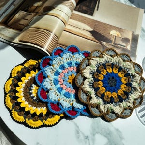 May include: Three handmade, crocheted coasters in a floral design. The coasters are in shades of yellow, brown, blue, red, and cream. They are arranged on a white surface, with a magazine visible in the background.