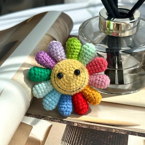 Peut inclure: Une fleur en crochet arc-en-ciel avec un visage souriant jaune au centre. La fleur est faite de fils de différentes couleurs, notamment du vert, du bleu, du rouge, du violet et du jaune.