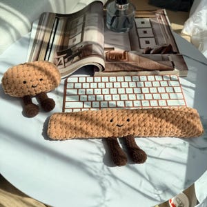 May include: Two handmade, light brown crochet items with smiling faces and dark brown legs. One is a long rectangle, the other is a mushroom shape. They are on a white surface with a keyboard and magazine in the background.